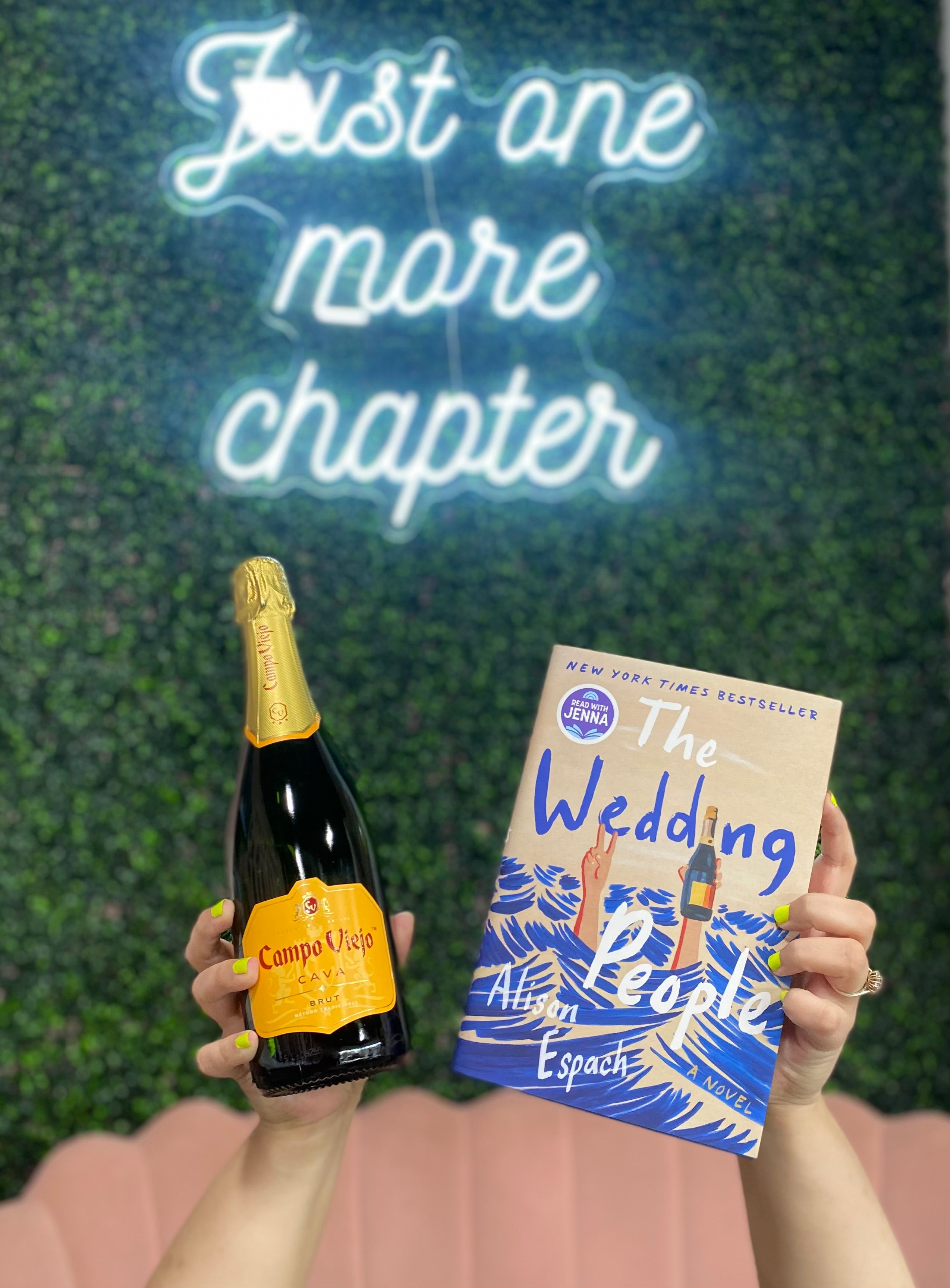 Bottle of Campo Viejo wine and book titled 'The Wedding of Pele' held against a green wall with 'Just one more chapter' neon sign.